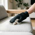 Hand sweeping a black mesh pet-hair cleaning mitt across a pale-gray fabric sofa, with a visible wad of cream cat fur lifted on the mesh